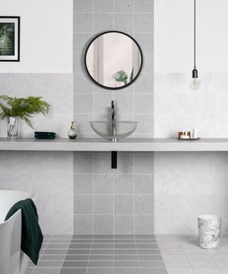 A gray tiled bathroom with counter, round mirror and clear glass sink.