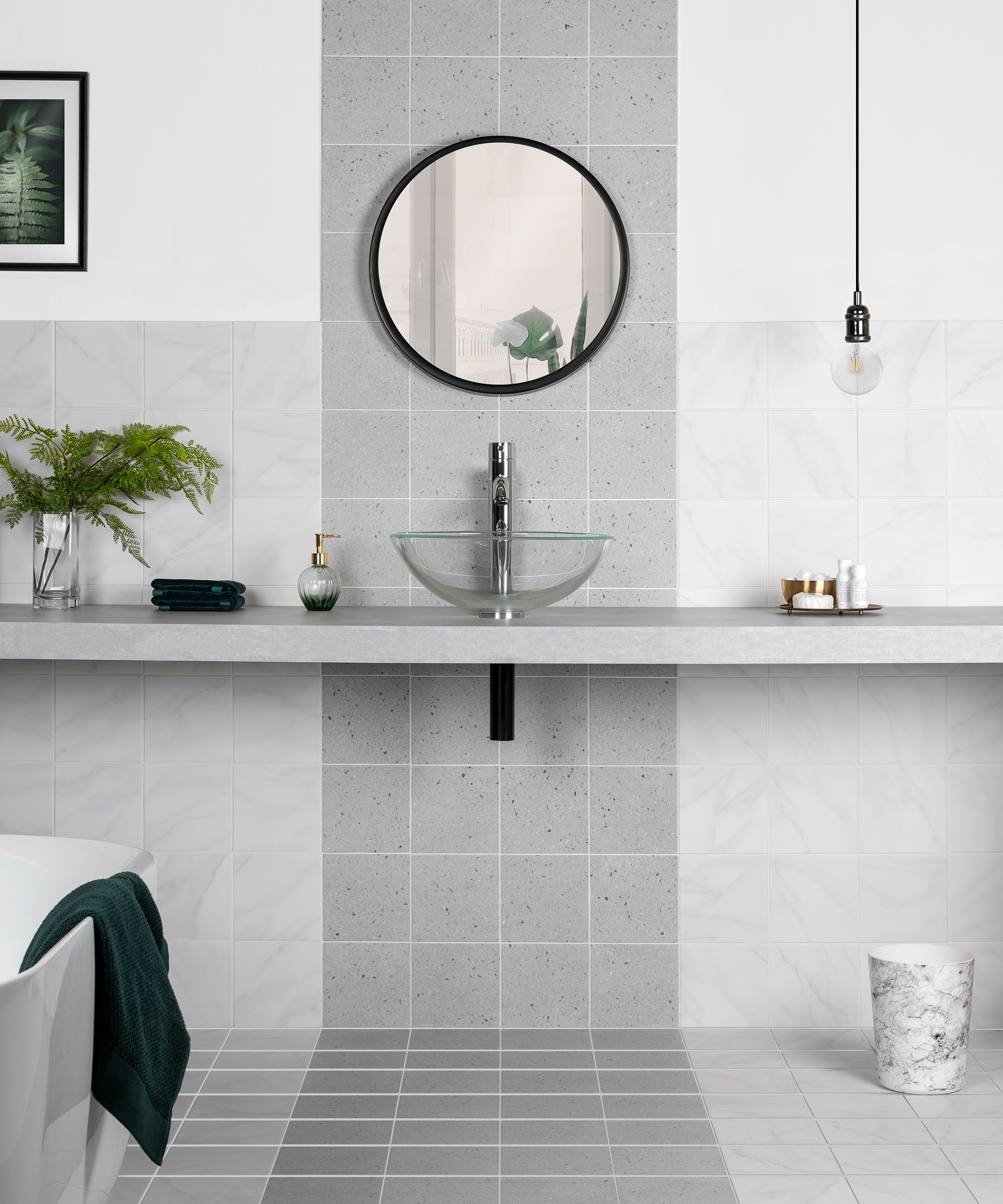 A gray tiled bathroom with counter, round mirror and clear glass sink.