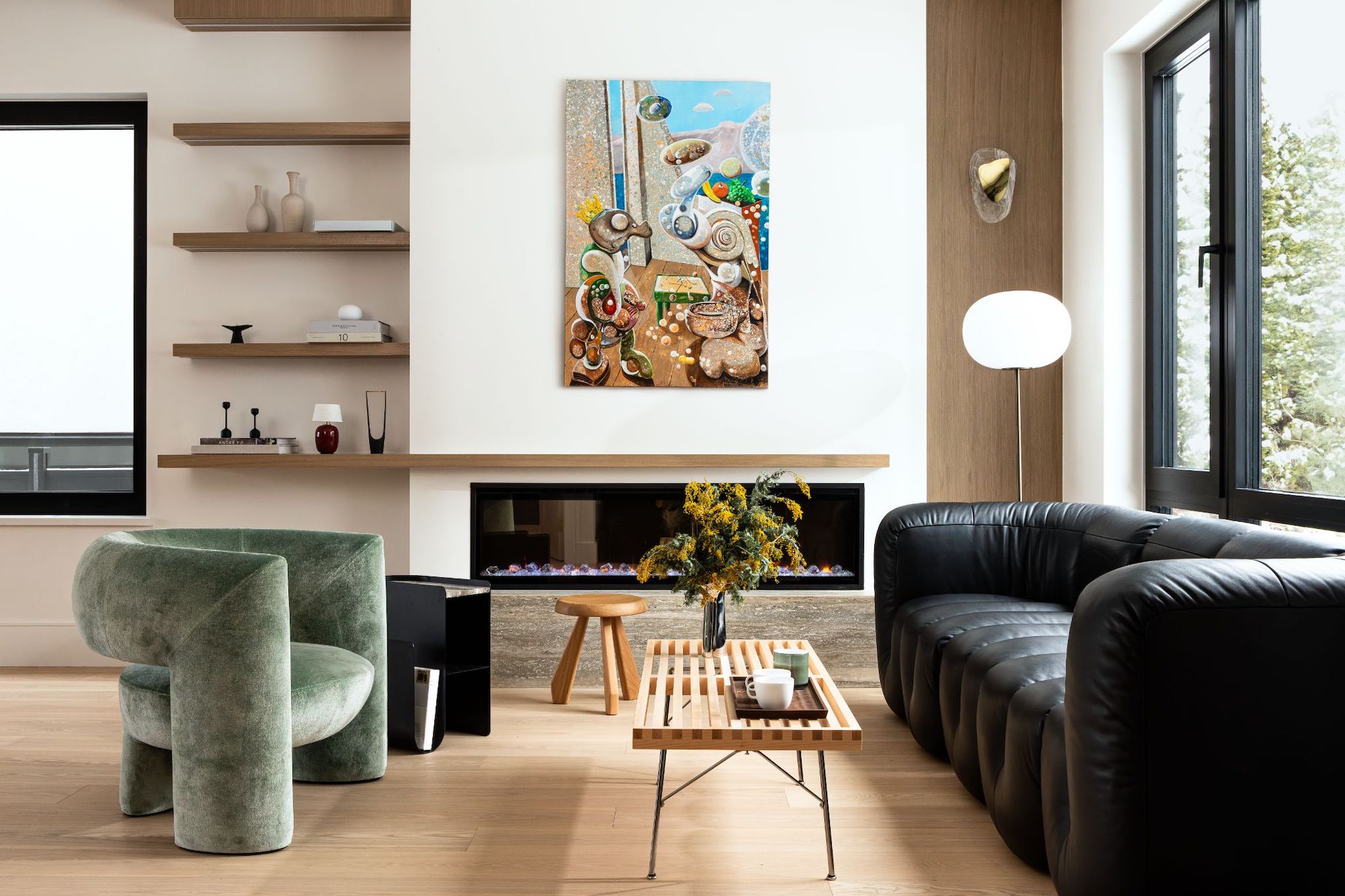 A view of the fireplace in the modern living area; white walls and light wood details, with a large, black leather sofa and a green armchair, and an artwork above the fireplace.