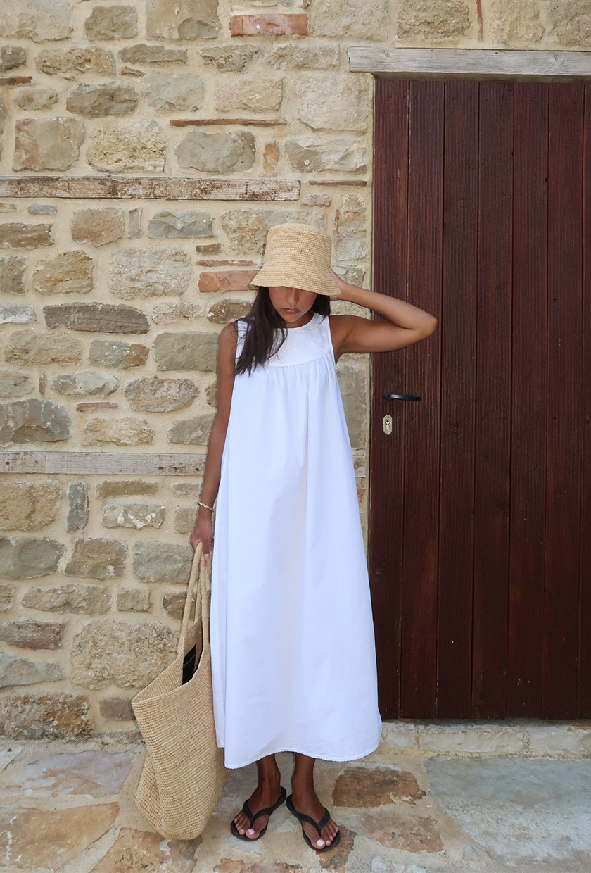 The summer aesthetic, coastal boho, is shown in a street style photo of a woman standing outside wearing a raffia bucket hat with a white maxi dress, black flip-flops, and an oversized raffia tote bag