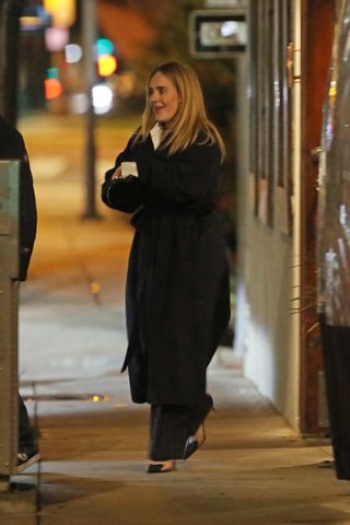 A photo of Adele pairing pinstripe pants with a black trench coat, a heart-shaped bag, and Alaïa naked pumps.