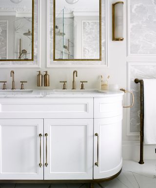 A close up shot of his and hers sinks with gold faucets and white cabinets with gold handles below two rectangular mirrors