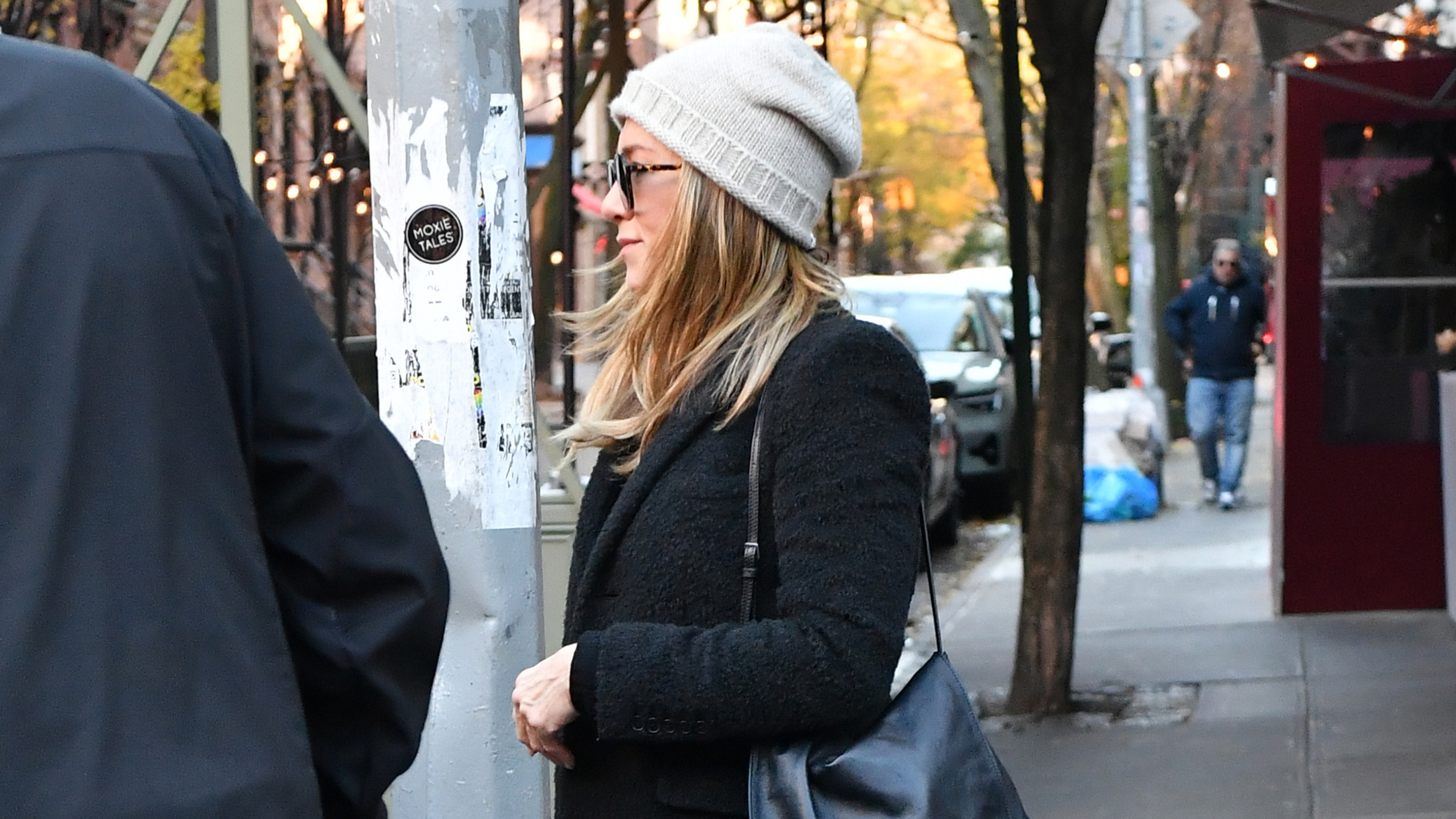 Jennifer Aniston in New York City wearing a black coat, tan hat, wide-leg jeans, and white sneakers.