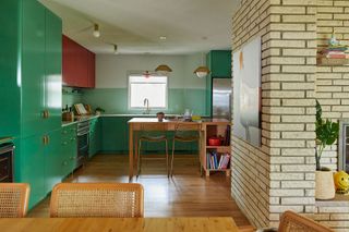 kitchen with green base units and red wall cabinets with wooden table cane chairs red-topped island pale green tiles and exposed brick wall