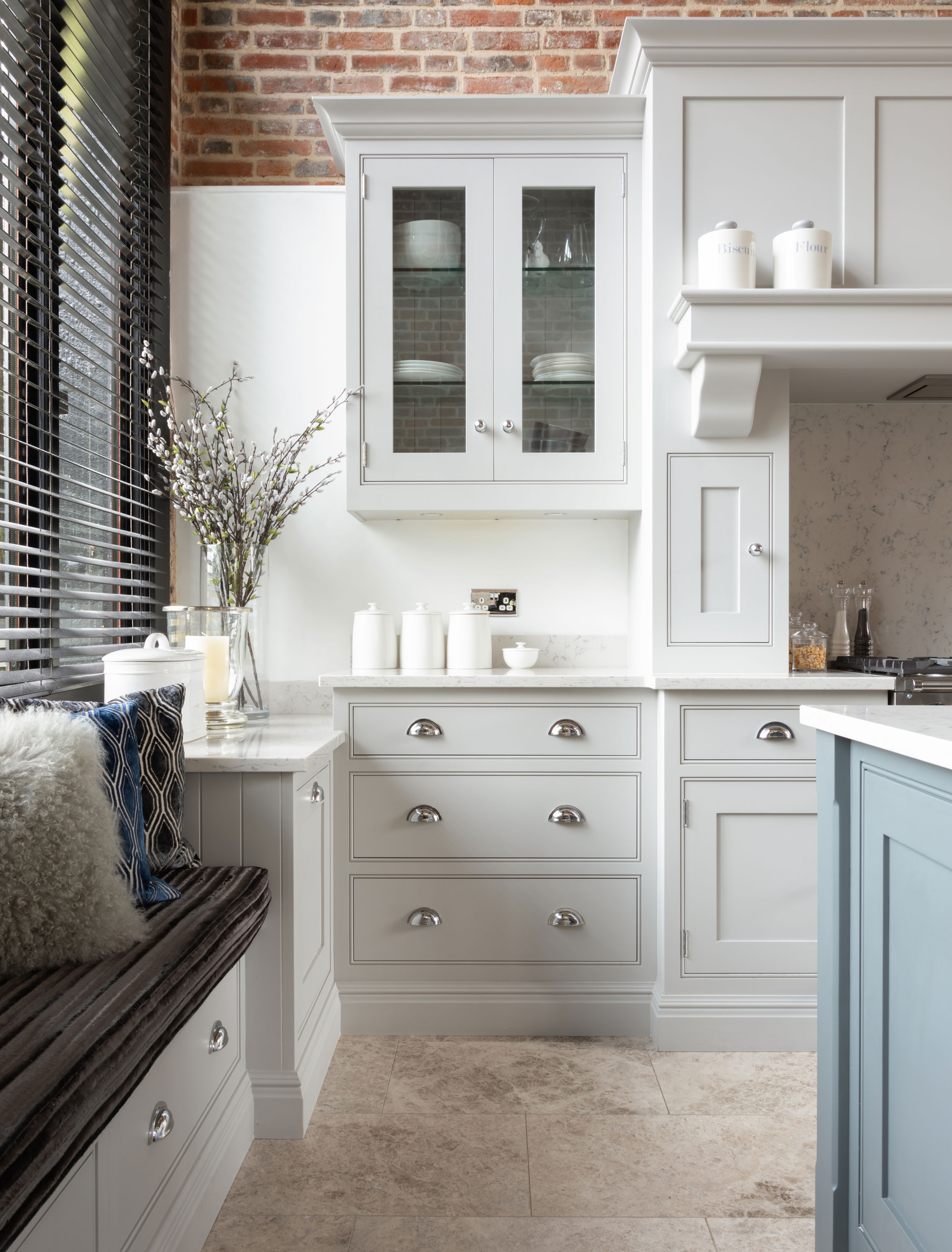 A kitchen with pale gray and gray-blue kitchen cabinet colors in a Shaker style, with stone flooring and exposed brick walls.