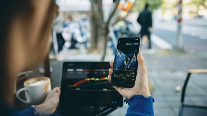 At an outdoor cafe, a woman sits with a smartphone and a laptop, both screens showing stock market charts.