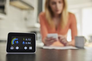 close up of a smart meter with woman calculating energy bills in the background