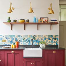 a kitchen countertop with dark red cabinets with brass cup pulls, and a striking blue graphic tiled splashback and a large Belfast sink with black tap