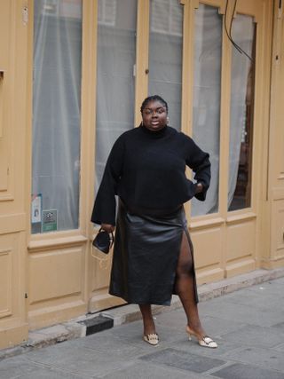 London-based fashion influencer Abisola Omole wearing a black sweater, black leather-look Mango skirt with a high split, and Manolo Blahnik mules and carrying a black mini bag.