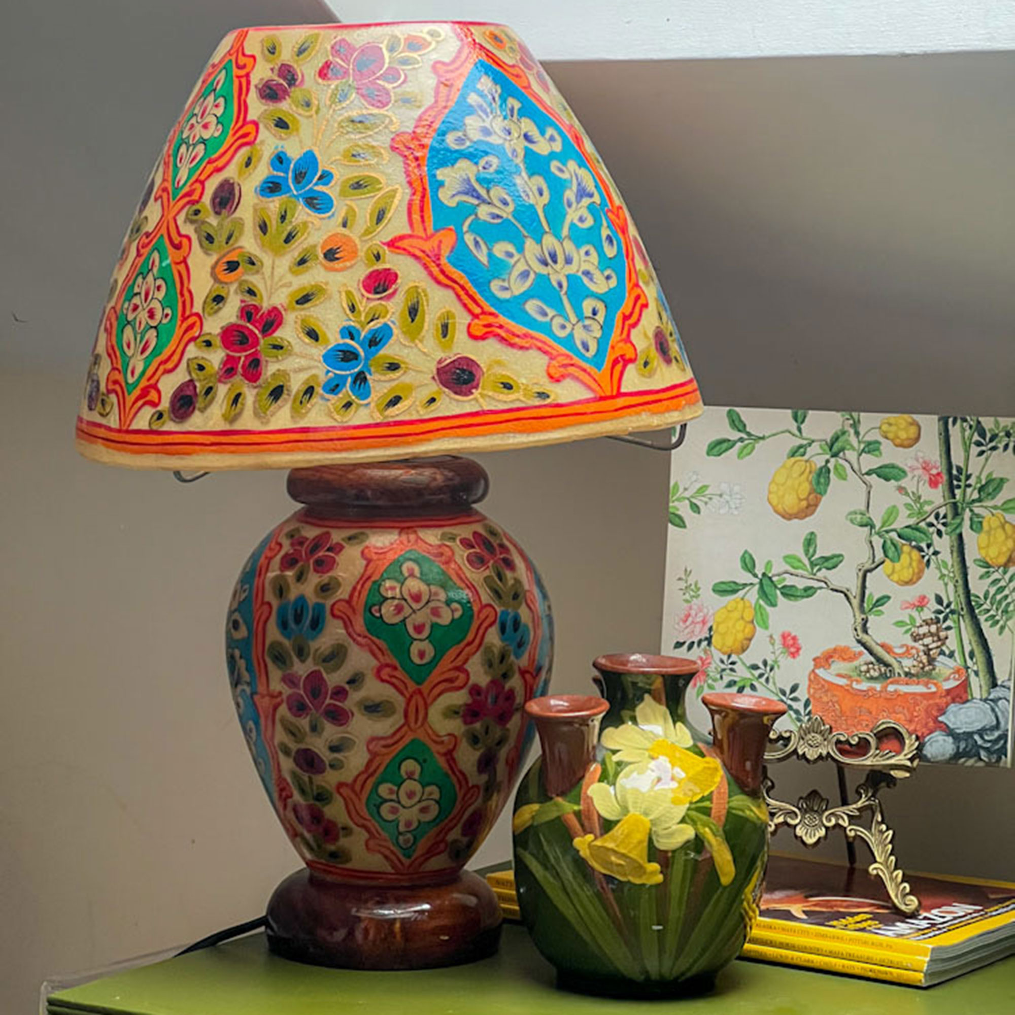 Lamp with colourful patterned base and shade beside small vase painted with daffodils and painting of lemon tree, all on side table