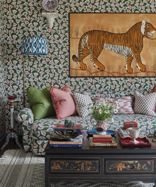 Lots of pattern in living room with wallpaper matching sofa and ikat lampshade and tiger artwork on wall and rug and antique style painted ottomana