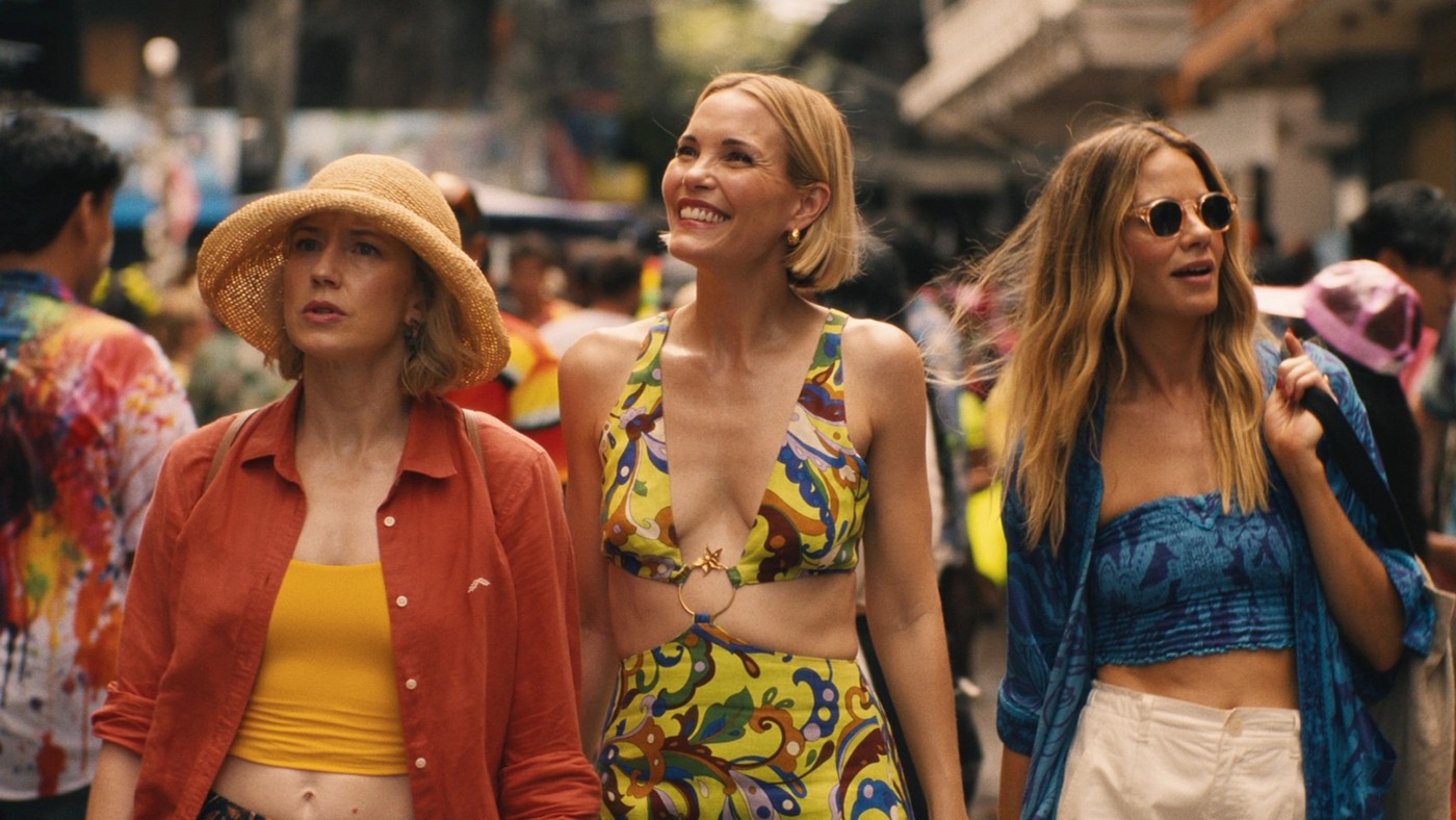 Scene from The White Lotus. Three stylish women walk through a bustling tropical market, dressed in colourful, summery clothes and looking curious, amused, or thoughtful.
