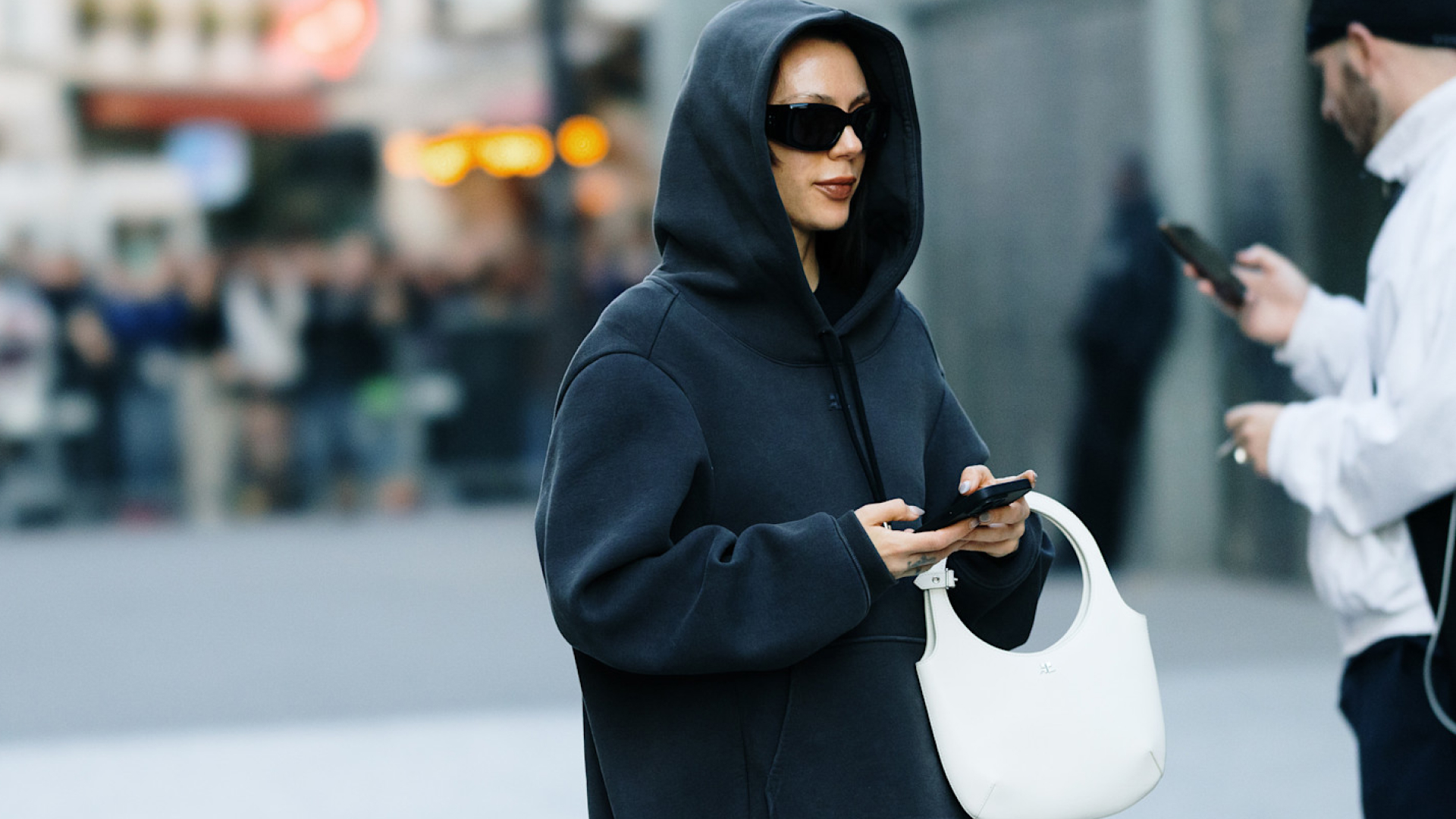 Fashion week attendee wearing black hoodie, black sunglasses, and white handbag