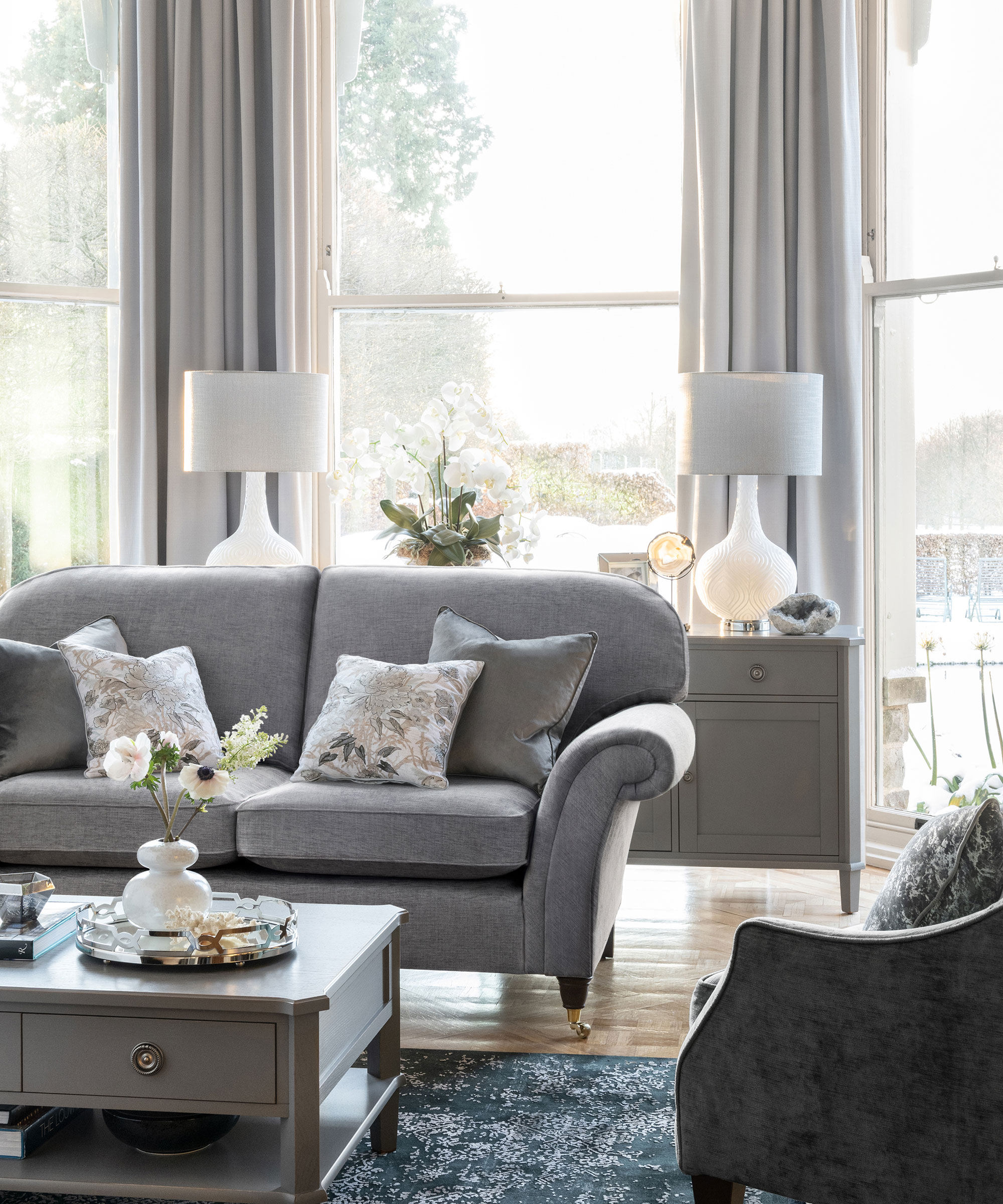 Gray sofa with patterned cushions in front of bay window with light gray curtains and gray cabinet with flowers and white/gray matching table lamps
