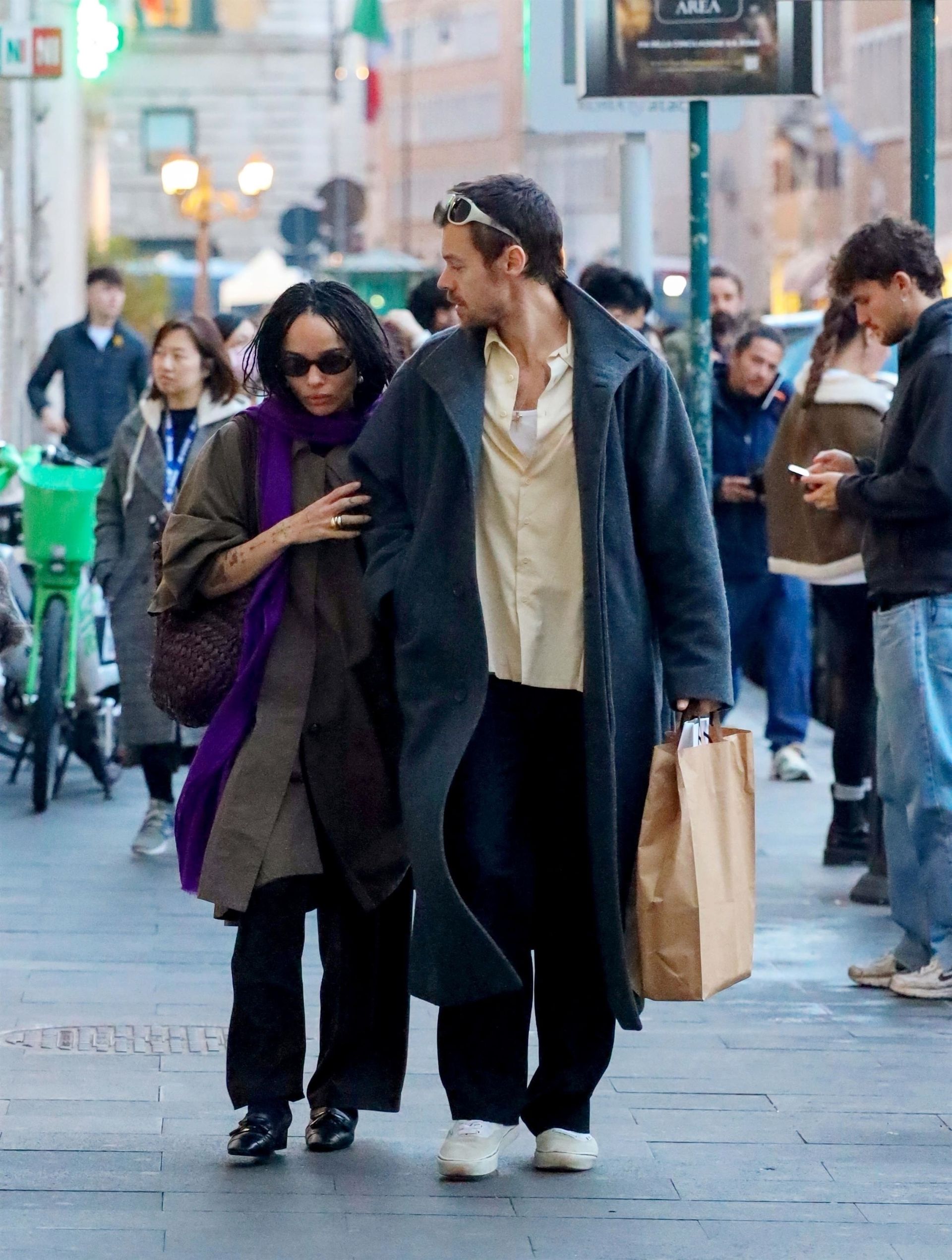 Zoe Kravitz walks down the street holding hands with Harry Styles. She wears a khaki trench coat with a purple scarf and patent shoes, whilst he wears a blue trend with a yellow shirt.