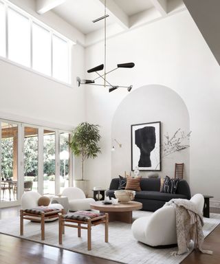 Organic modern living room with white walls and high ceilings. Large windows allow natural light to flood the space, white house plants bring life to the space. Large cozy sofas add comfort, and an organic shaped wooden coffee table centres the scheme