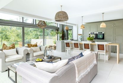 Open plan kitchen in neutral colors with rattan style sofas and chairs and gray green cabinets and picture windows