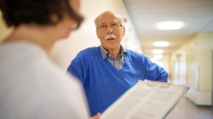 A man talks with a nurse holding a clipboard in a hospital hallway.