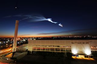 A SpaceX Falcon rocket soars into space in this stunning view from SpaceX's headquarters in Hawthorne, California. A used Falcon 9 first stage stands in the foreground.