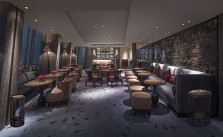 A bar area in the Shangri-La at the Shard hotel. A light gray couch sits against the wall to the right, with round tables in between the couch and light brown tabourets. Against the floor-to-ceiling windows, we have another row of sitting areas in the same fashion. The bar is to the center, up against the far wall.