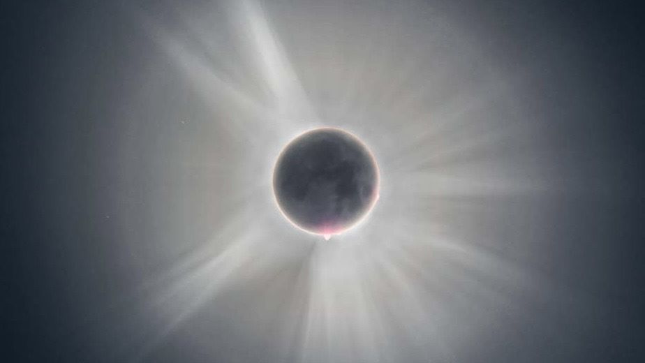 The moon&#039;s features are faintly visible. It is fully eclipsing the sun, which is hidden except for its corona glowing in white.