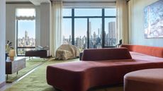 A modern living room with a sweeping view of Central Park with a sculptural red sofa and green carpet, a piece of art on the wall and wooden side tables
