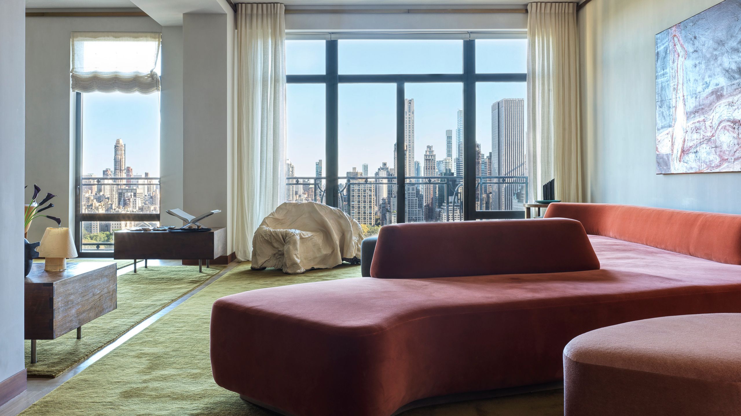 A modern living room with a sweeping view of Central Park with a sculptural red sofa and green carpet, a piece of art on the wall and wooden side tables