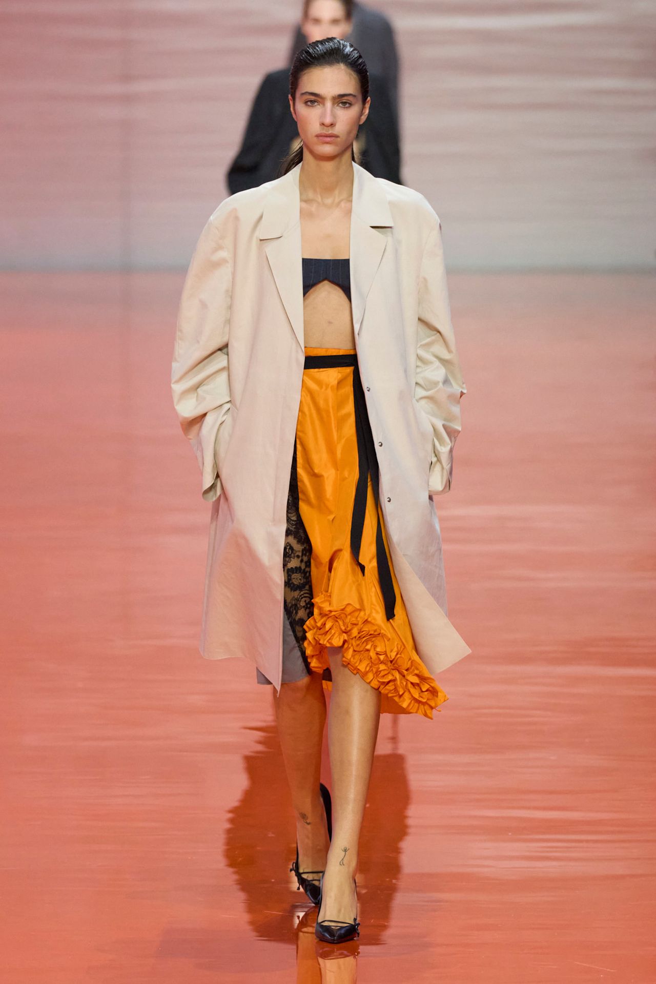 A Prada model wearing a light tan coat layered on top of a black, a tangerine orange midi skirt with a ruffled hem, a black belt, and a pair of black pointed kitten heels.