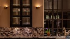 Two sconce lights are shining on the wall of a kitchen at night. There is a cabinet on the wall and red and white marble countertops.