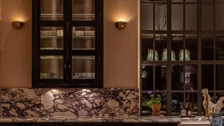 Two sconce lights are shining on the wall of a kitchen at night. There is a cabinet on the wall and red and white marble countertops.