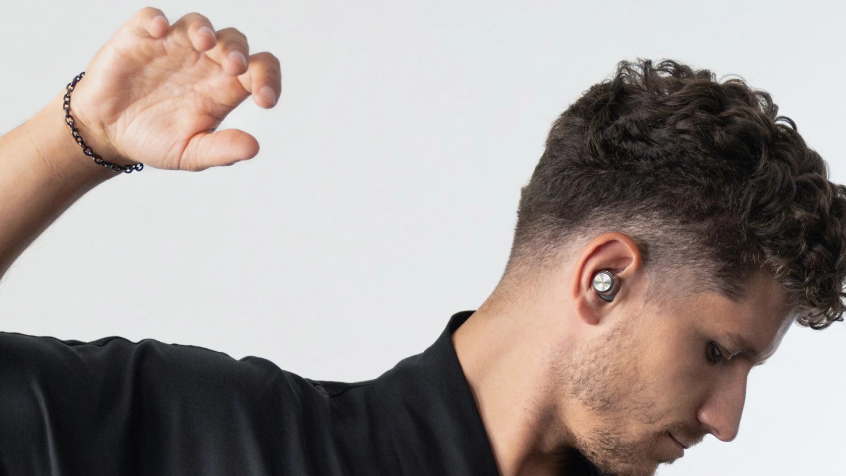A profile shot of a man with short, dark, curly hair and a slight beard, wearing a .Technics EAH-AZ100 wireless earbud in his right ear and raising his left arm, set against a bright white background