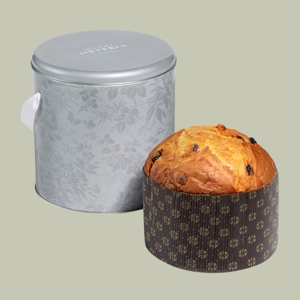 A silver tin decorated with floral motifs and with a white satin handle, placed next to a panettone on a pale green background.