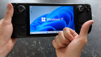 A Steam Deck running Windows 11 is held in both hands, with the operating system displayed prominently on the screen while a thumbs-up gesture partially covers the lower-right corner.