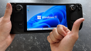 A Steam Deck running Windows 11 is held in both hands, with the operating system displayed prominently on the screen while a thumbs-up gesture partially covers the lower-right corner.