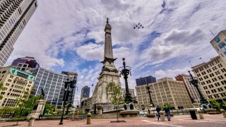 Six jets in formation fly through a cloudy blue sky over a city's center square, a tall monument tower standing in the middle with building surrounding. 