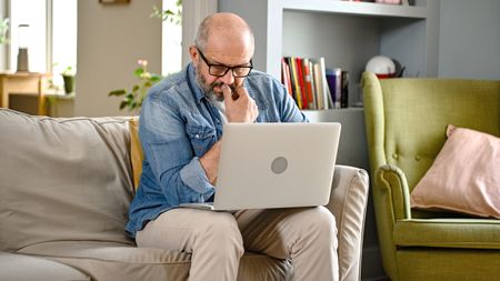 An older man stares intently at his laptop while sitting on his sofa.
