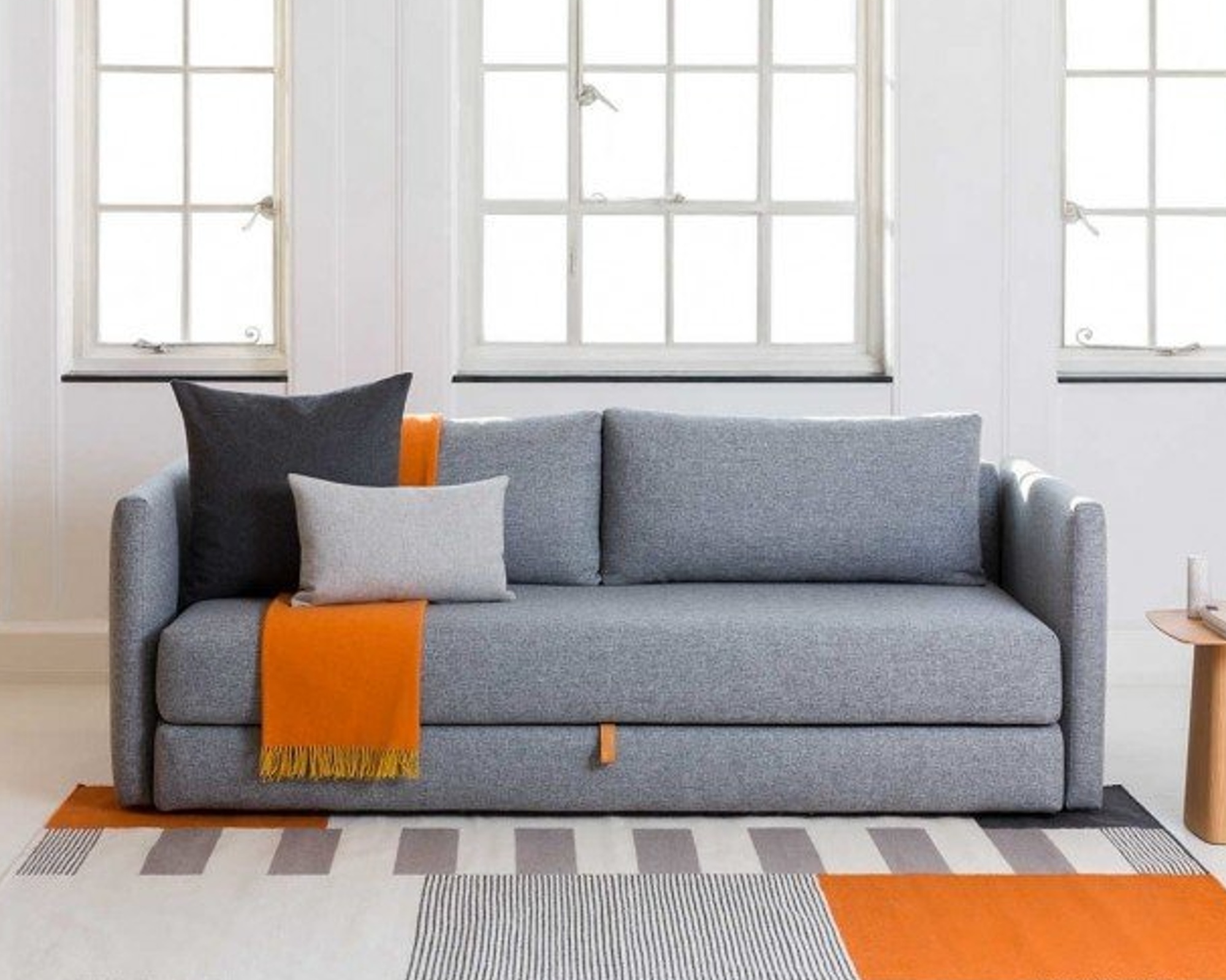 A grey sofa bed sat on a geometric orange patterned rug in front of three brightly lit living room windows