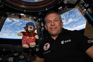 a man in a black polo shirt poses with a small plush bear near a window of the international space station, with earth visible in the background.