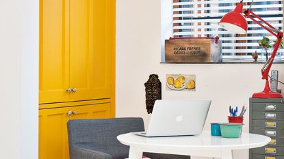 laptop on white table in kitchen with yellow cupboard door and red table lamp