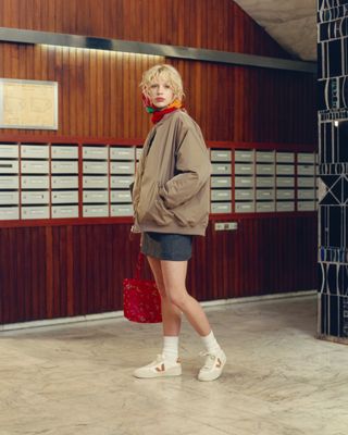 A model wears navy shorts, a tan jacket, and white Veja sneakers.