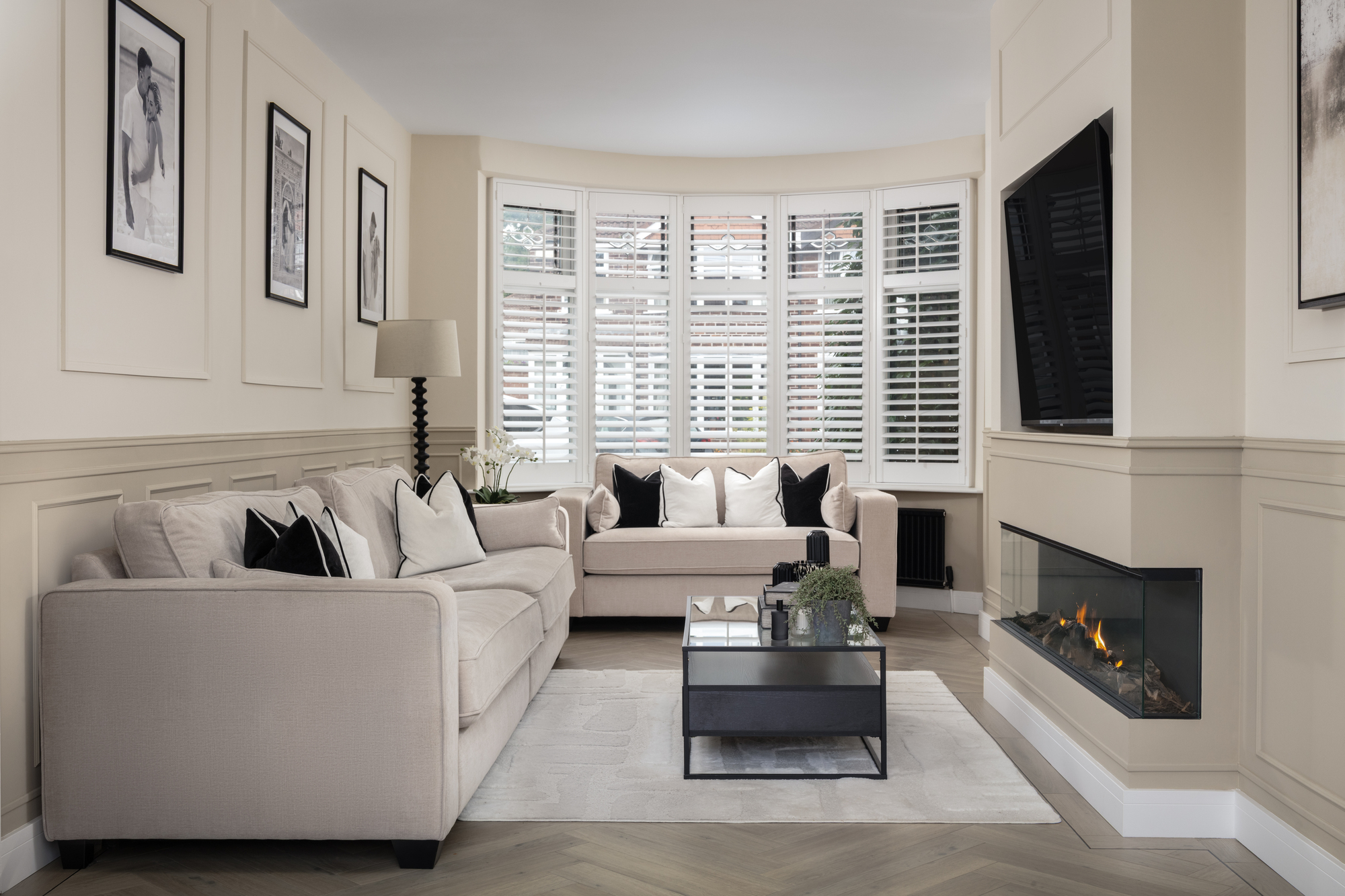 A small neutral living room with wainscoting, a bay window, and a TV above a fireplace