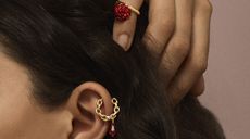 A close-up of an ear with a small gold loop chain ear-ring dangling from the top outer part with a red diamond-shaped pendant. Above the ear is a hand with a gold-coloured ring with red flower shaped pendant on the outer finger. 
