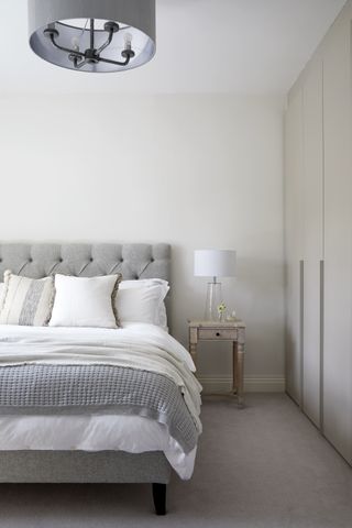 grey and white bedroom with grey button backed bed, antique side table, white lamp, fitted wardrobes, grey pendant