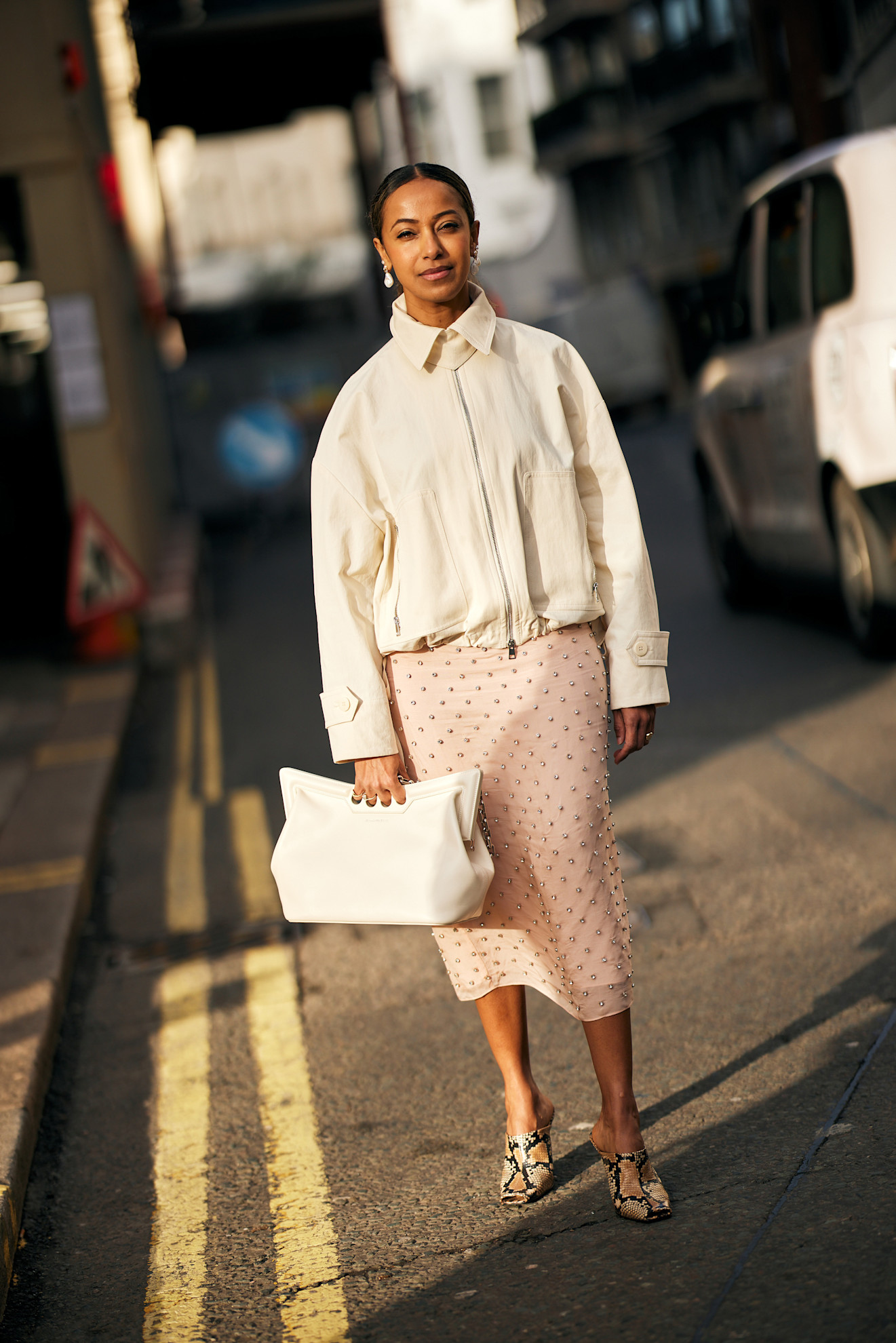 a woman at London fashion week wearing a white jacket, pink studded skirt, and snakeskin mules