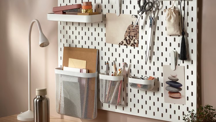 White IKEA SK&amp;Aring;DIS pegboard mounted on a wall above a desk with shelves, containers, hooks and a letter holder holding stationery and accessories