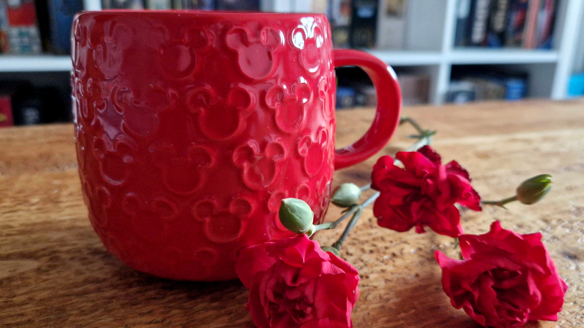 A red mug with Mickey Mouse icons on it beside roses laid out on a wooden table 
