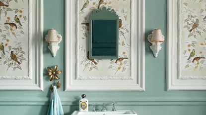 Small bathroom wall paneling ideas look so smart. Here is a bathroom with white wall panels with brown bird wallpaper, teal walls, two white wall sconces, a white standing basin, and a light blue towel