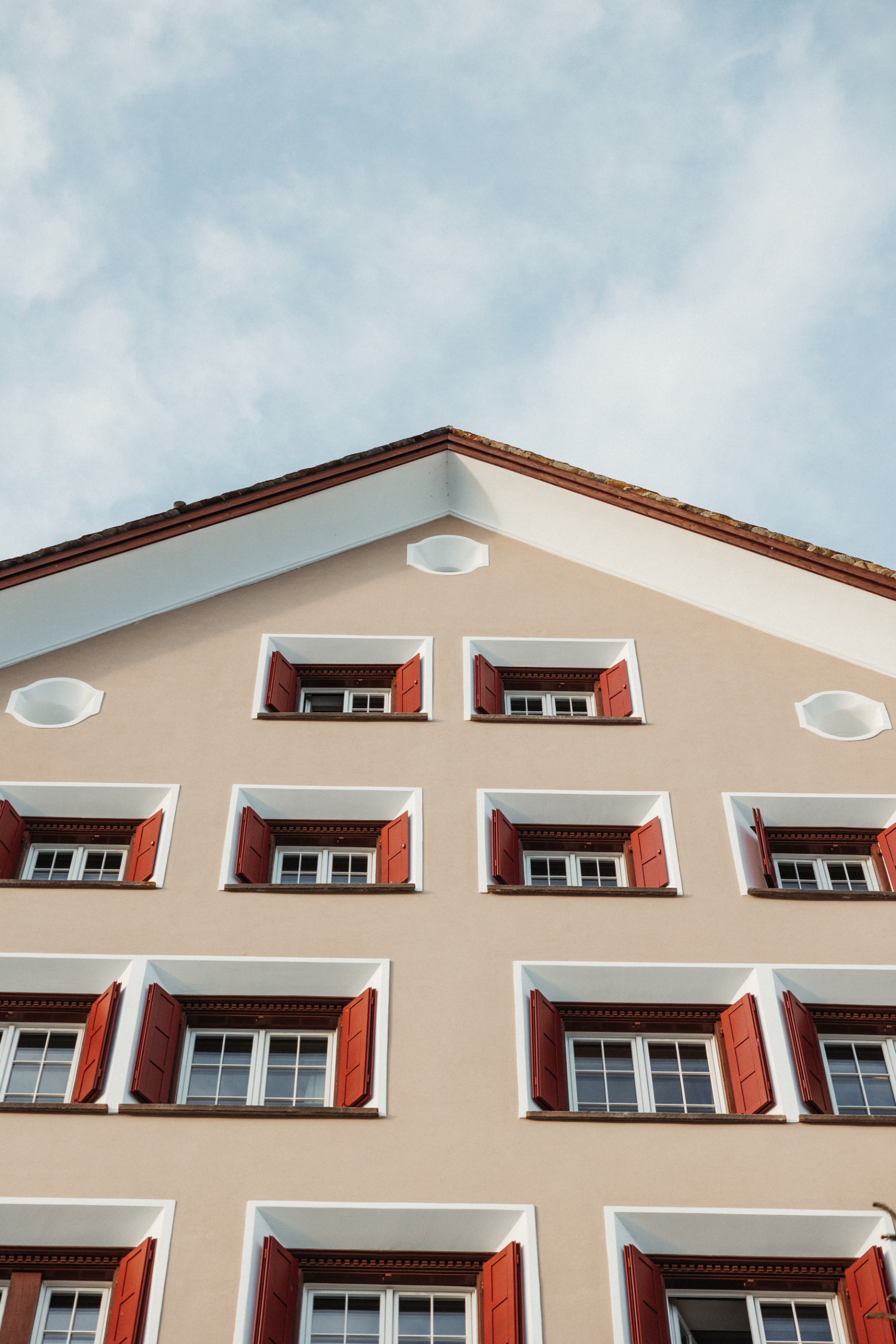 A Wes Anderson-style photograph of a building's facade. It has beige walls and white windows with red shutters