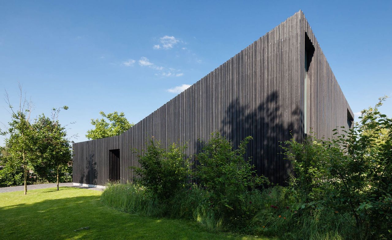 The exterior of a residential home clad in a dark, heat-treated wood with glass doors, surrounded by green mowed lawn and trees and a view of the skyline