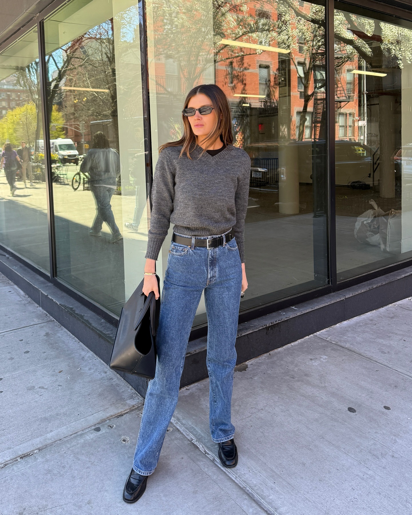 NYC style influencer Coco Schiffer poses in a gray V-neck sweater layered over a black T-shirt, black belt, bootcut jeans, a black minimal tote bag, and black loafers